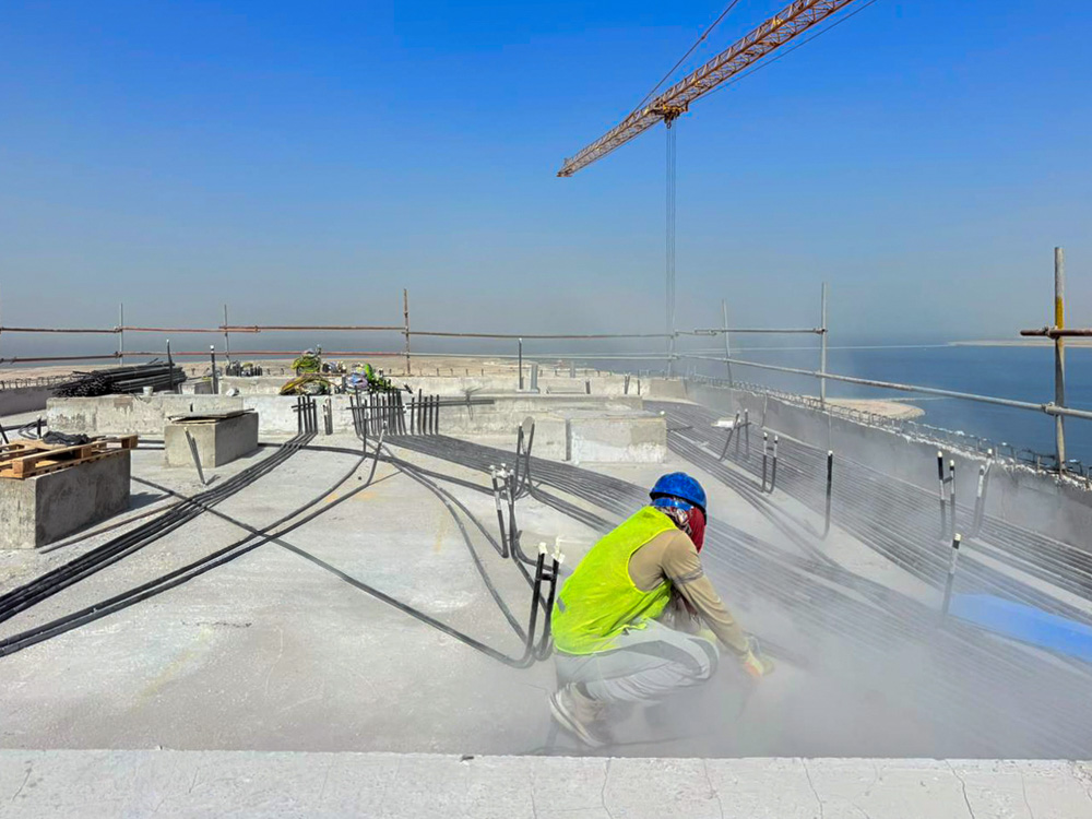 Beach Walk Iii By Imtiaz A construction worker in a yellow vest kneels on a rooftop with machinery and cables, overlooking a water body.